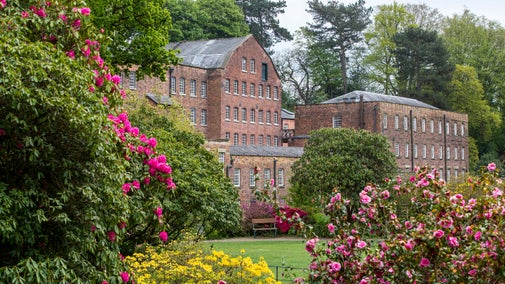 A large brick building with many windows, set in a garden of colourful shrubs and backed by leafy trees